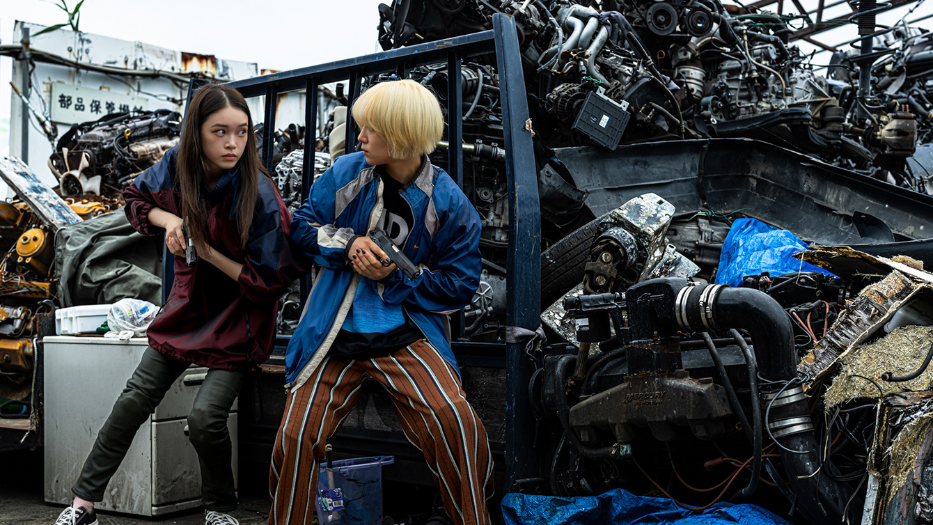 Chisato and Mahiro look intense, guns drawn, at the entrance to a scrapyard