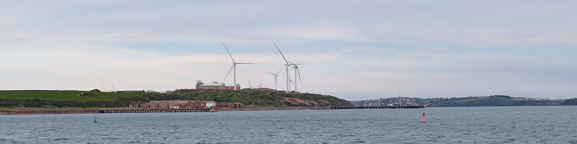 Some gas tanks and wind turbines are visible across a body of water. This is Dragon LNG terminal, or what can be seen from Milford Haven